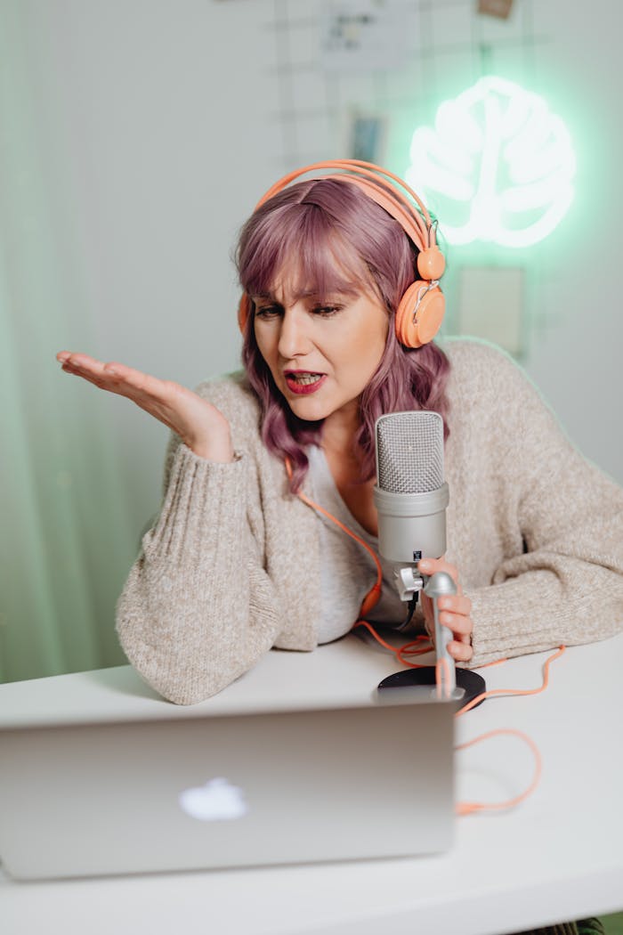 A woman using a microphone and headphones, speaking into a laptop during a podcast recording session.